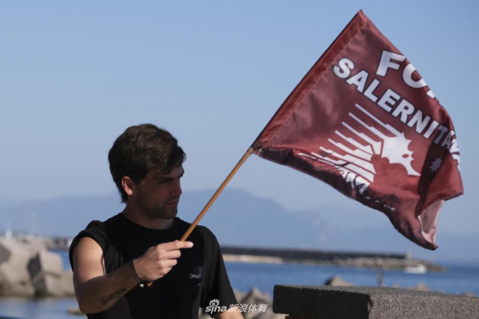  一辆被球迷簇拥的汽车车顶，两名男子激动地探出身子，手中紧紧握着一件巨大的萨勒尼塔纳队旗。背景中，整条街道变成了红白两色的河流，无数面旗帜在风中猎猎作响，描绘出小城足球最原始也最动人的庆祝画面。