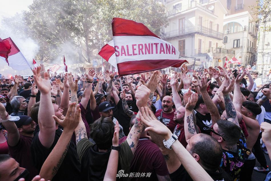  一名年轻的萨勒尼塔纳球迷难掩激动之情，在狂欢的人群中高高挥舞着巨大的红白色队旗，旗帜上标志性的海马图案在夕阳和烟雾中迎风飘扬，仿佛在向整个城市宣告王者的归来。