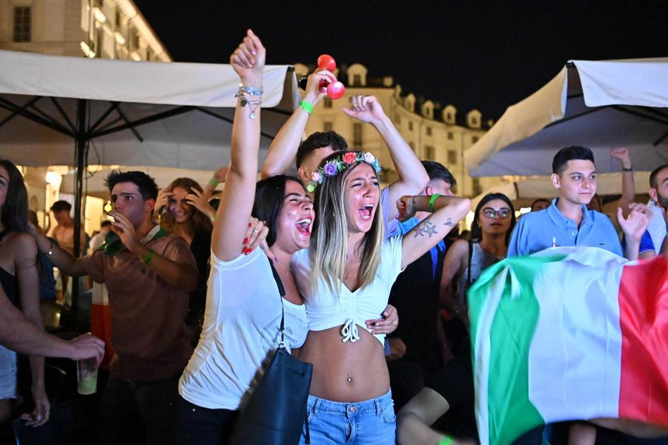 一名年轻的意大利球迷骑在朋友的肩膀上，双手高举过头顶竖起象征胜利的食指，脸上涂着绿白红三色油彩，在人群的簇拥中对着镜头忘情欢呼。