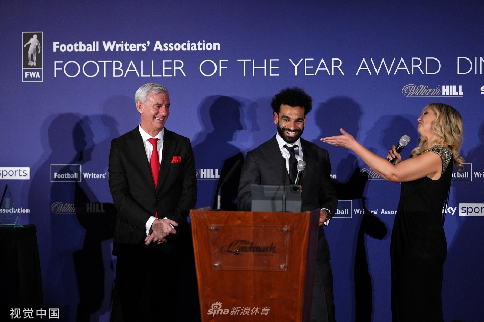 颁奖典礼现场，萨拉赫手持奖杯向在场的观众与媒体致意，背景是隆重的晚会布置，气氛热烈而庄重。