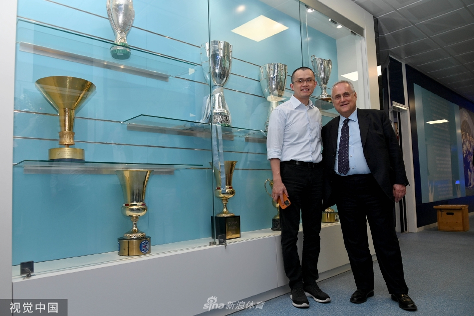 赵长鹏手持属于自己的天蓝色拉齐奥球衣，与球队队长亲切握手，背景中是绿茵场和训练的球员