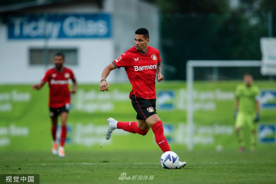 勒沃库森球员进球后张开双臂奔向角旗区庆祝，身后队友激动跃起，远处落日余晖将球场染成金色