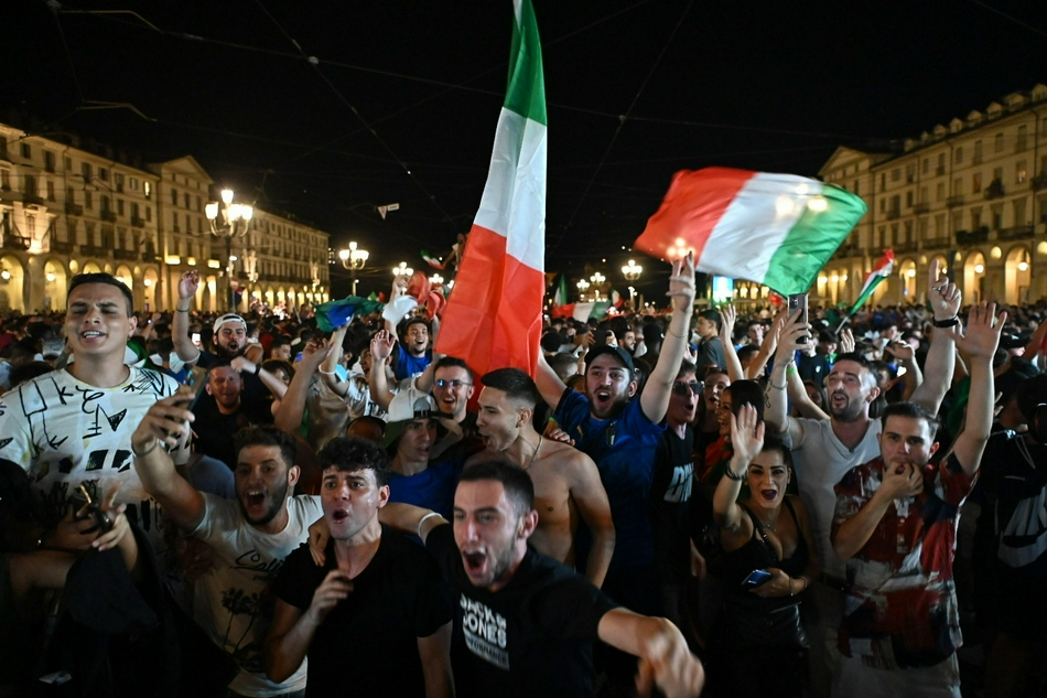 在都灵胜利广场的纪念碑下，一对情侣相拥而吻，背景是漫天飞舞的纸屑和欢呼雀跃的人群，他们的脸上洋溢着幸福与自豪的笑容。