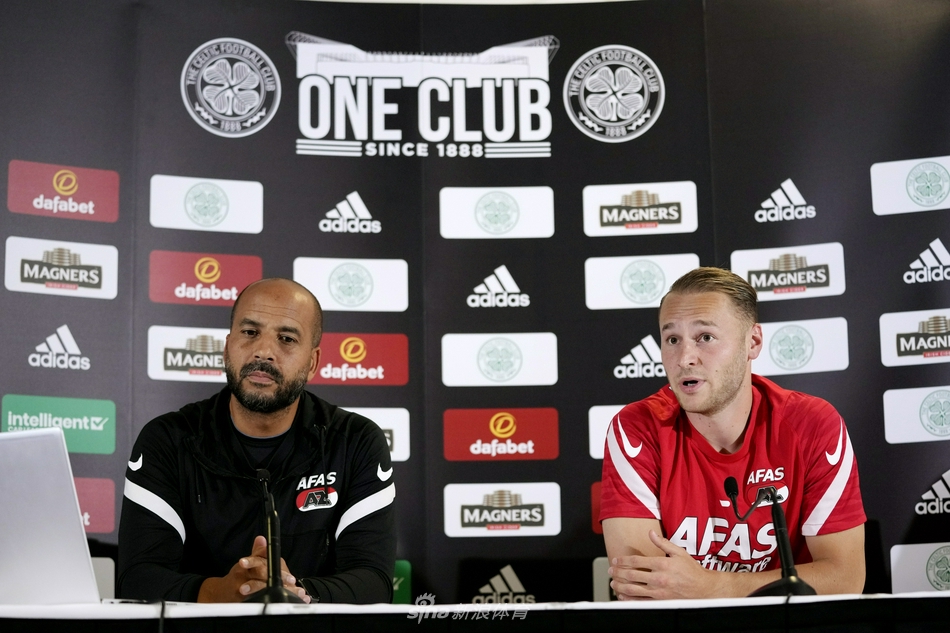 延森教练在发布会中神情专注地倾听记者提问，微微前倾的身体姿态显示出他对这次欧战征程的重视，桌上摆放的矿泉水瓶和麦克风见证了这场赛前的智慧博弈。