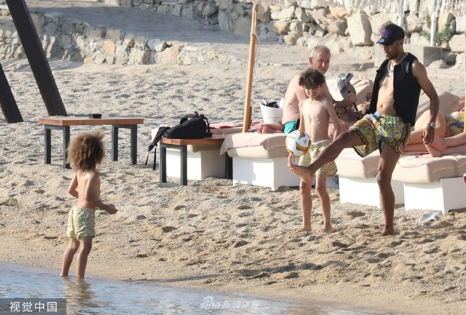 奥巴梅扬在沙滩上躬身指导儿子控球，远处湛蓝的爱琴海与他们的专注神情形成宁静而温馨的对比