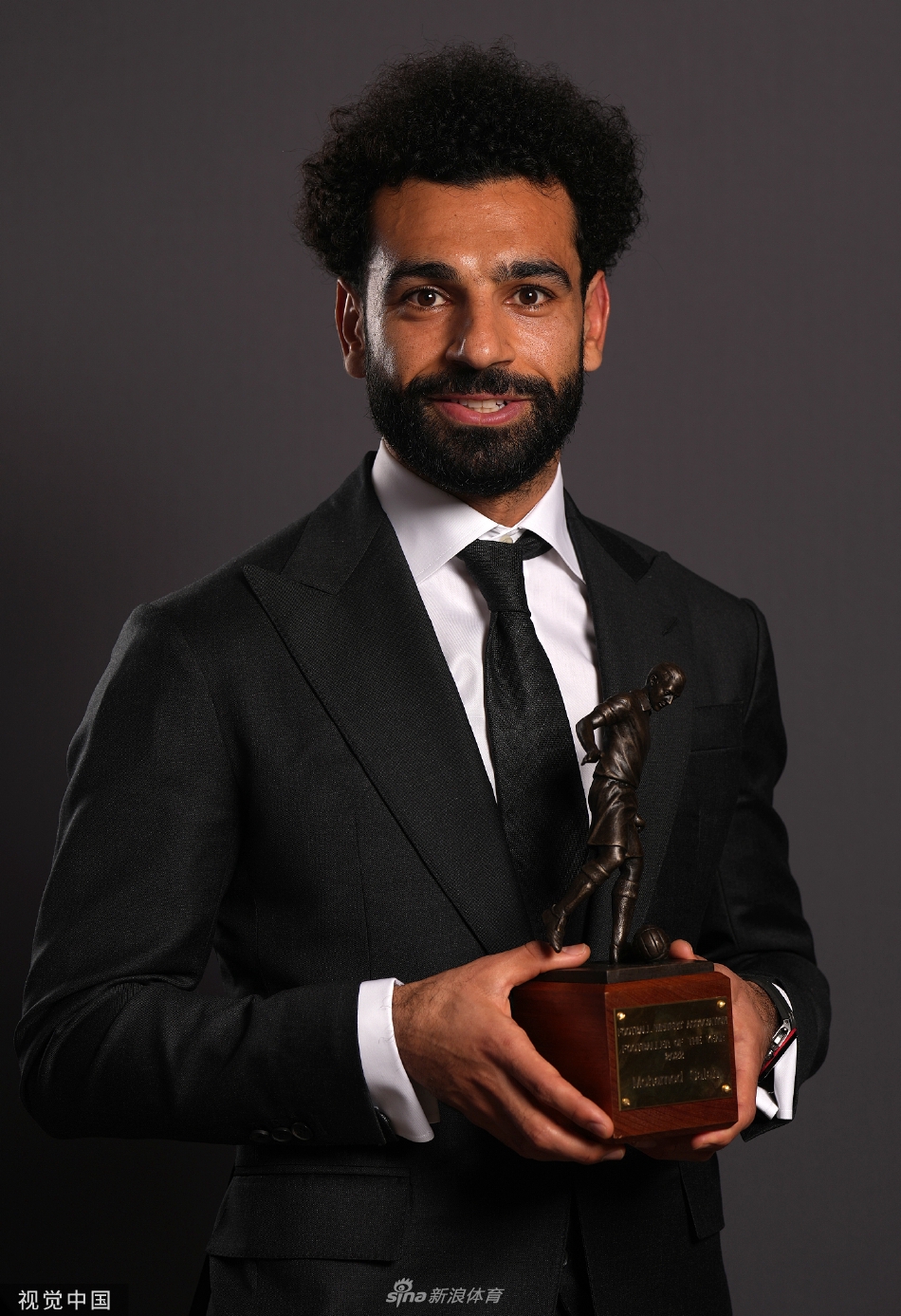 萨拉赫身穿优雅正装，在镜头前自信地展示着手中的年度最佳球员奖杯，展现出世界级球星的从容风范。