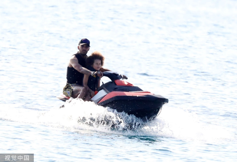 奥巴梅扬轻松地用膝盖颠球，儿子尝试模仿，背景中的遮阳伞与沙滩椅点缀着悠闲的假日气氛