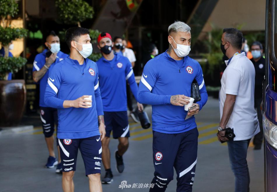 镜头拉近，捕捉到了更加触动人心的细节。一位智利球员正弯腰拾起地上的训练装备，这一简单的动作却被赋予了别样的仪式感。他的侧脸写满了专注与风霜，那是无数次拼抢、无数次冲刺后留下的岁月印记。身后的队友三三两两交谈着，他们的笑声与不远处球迷的欢呼声交织在一起，构成了大战前难得的宁静序曲。阳光透过枝叶的缝隙，洒在他们红色的战袍上，斑驳的光点仿佛是为即将出征的勇士们披上的祝福铠甲。