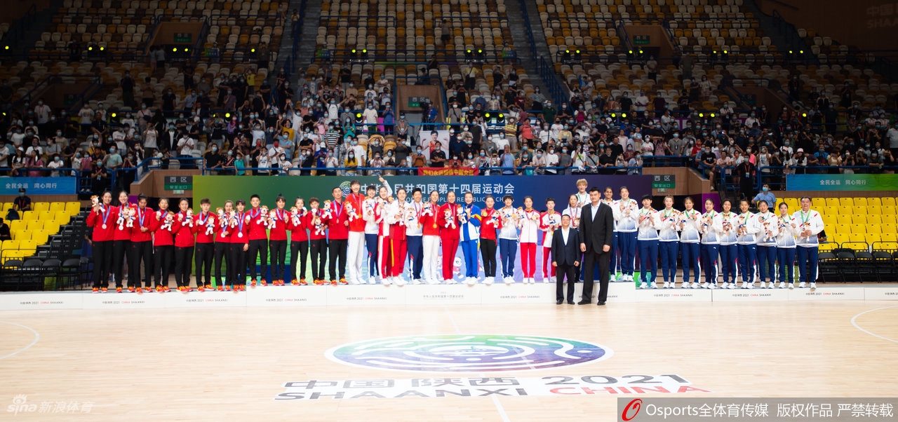 赛场全景图,红蓝色的地板映衬着激烈的对抗。江苏队球员正试图突破联合队的多人防线,虽然实力悬殊,但她们每一次进攻都全力以赴,毫不畏惧。