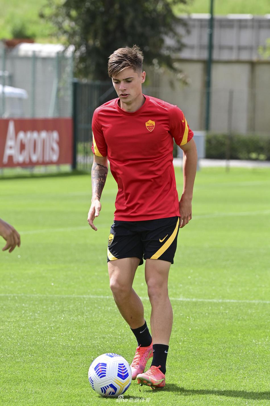 一名罗马球员正在进行带球绕杆练习，他低头专注控球，身体重心压低，皮球仿佛黏在脚下，周围摆放着橙色标志盘。