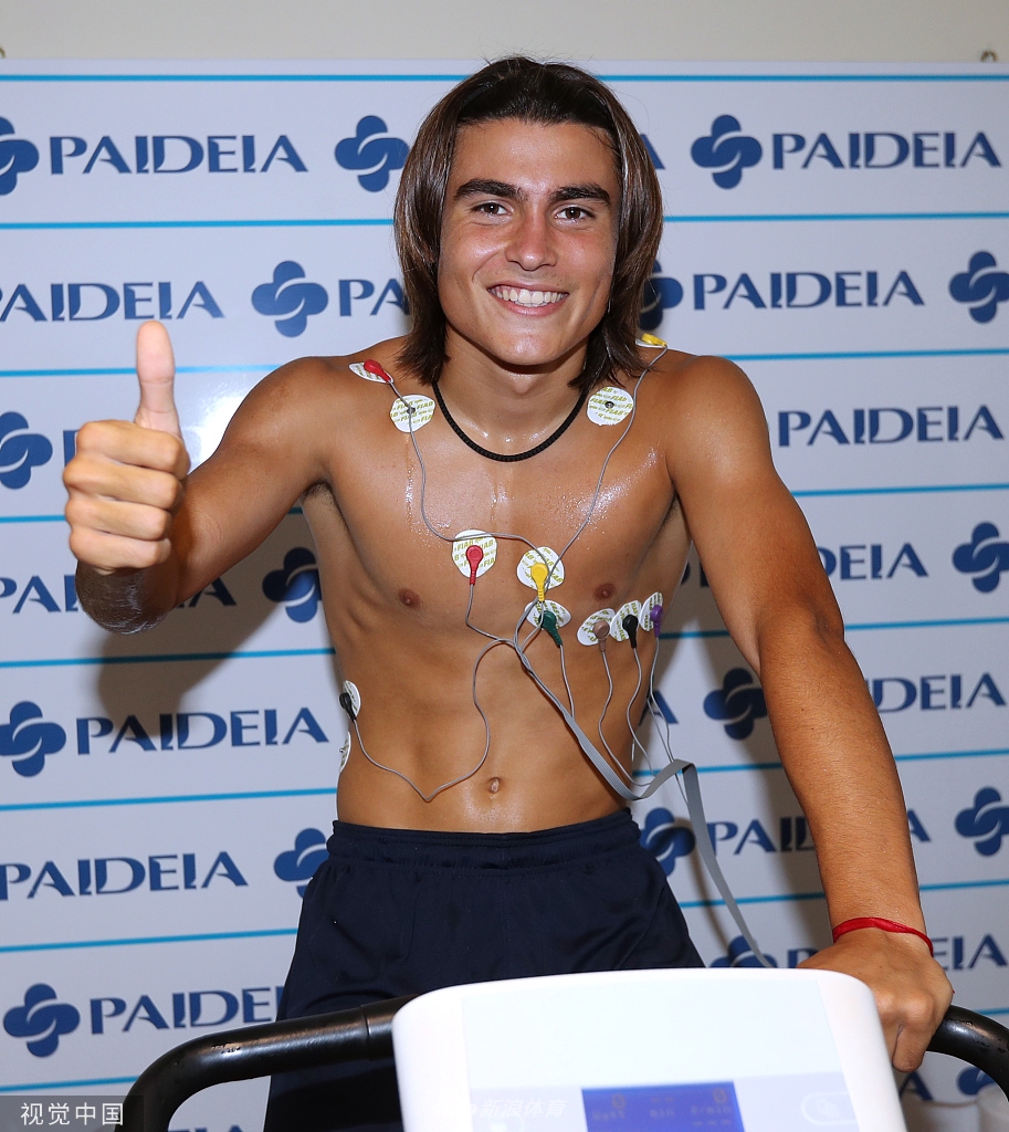 体检过程中的一个小插曲，卢卡·罗梅罗低头查看手机，似乎在向家人或经纪人报平安，身后是现代化的医疗设备，记录着这位足坛神童的身体数据。