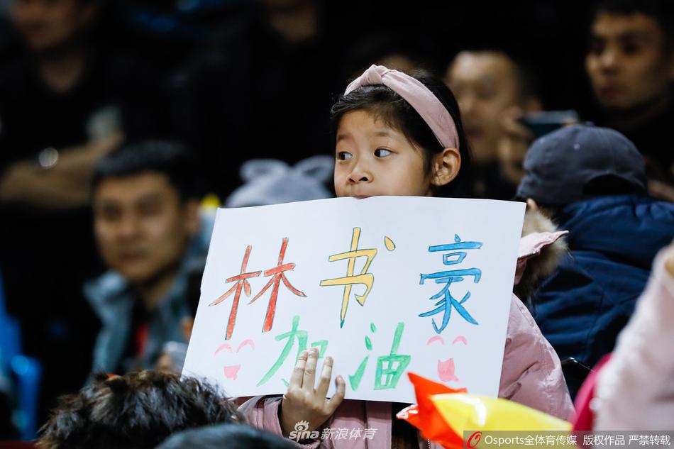 一名清秀的北京女球迷对着手机镜头俏皮地比出胜利手势，她身穿白色的北京首钢T恤，在周围吉林球迷的注视下，显得格外自信与亮眼。