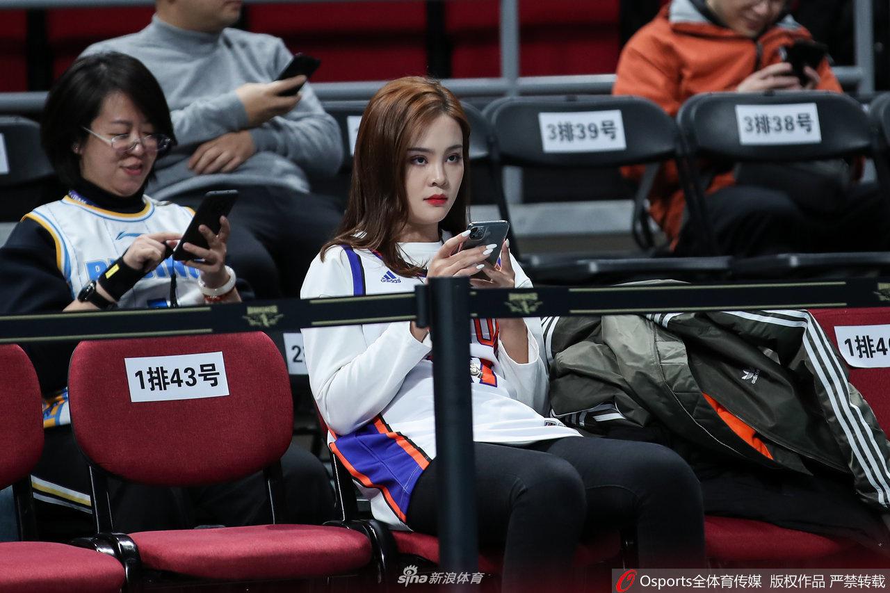 一位长相甜美的北京女球迷,利用暂停时间低头查看手机,似乎在与朋友分享此刻紧张的心情,她身旁的座位上放着助威用的充气棒,背景是座无虚席的沸腾球场。