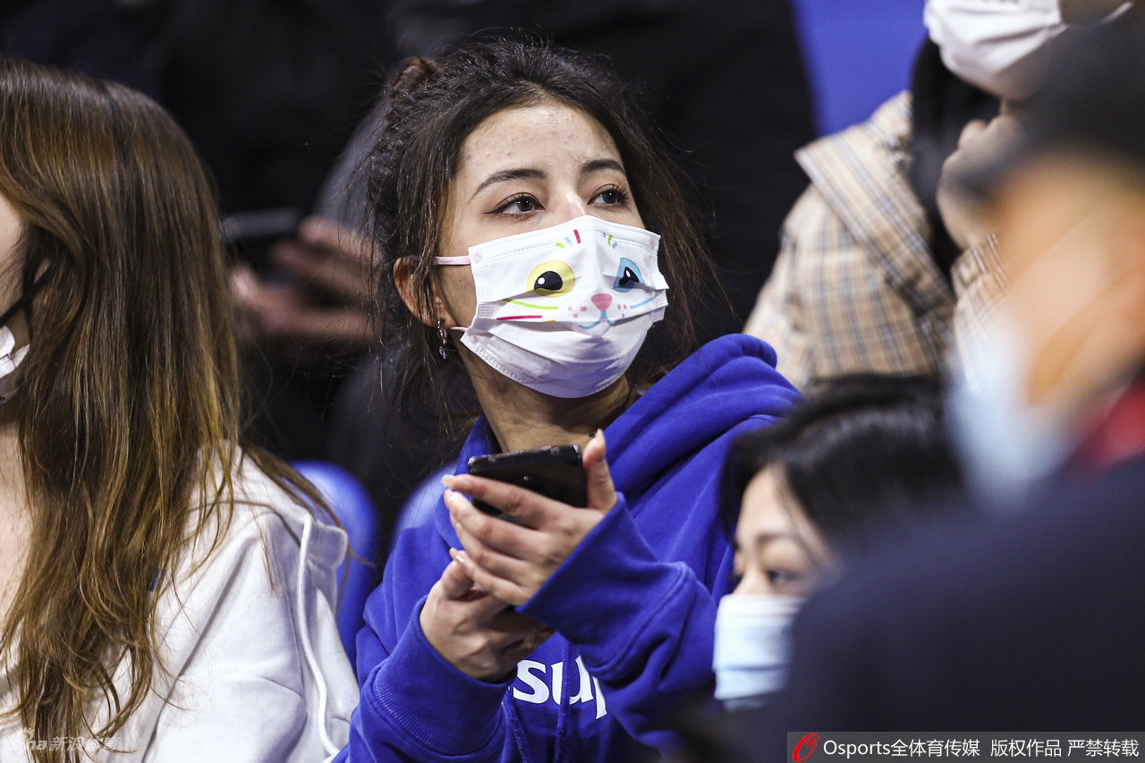 一位穿着浅色毛绒外套的长发女球迷，站在看台过道中回眸一笑，身后的红色座椅与她的温暖笑容形成了冬日里最治愈的反差，仿佛她的目光所及之处，正是赛场上最精彩的瞬间。