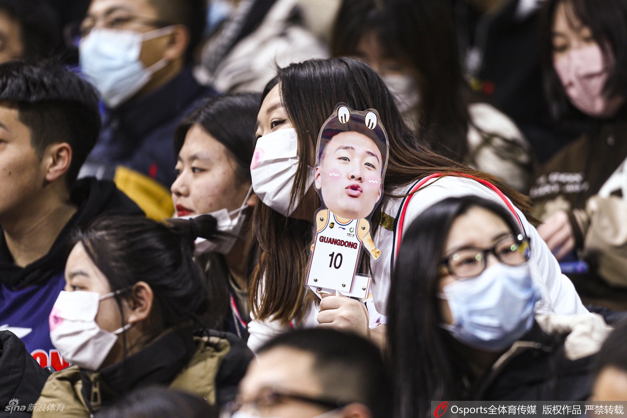 比赛暂停间隙，看台上的气氛依然热烈，这位戴着黑框眼镜的女球迷侧身看向镜头，背景里是正在布置战术的教练组和密密麻麻的观众，构成了一幅生动的赛场众生相。