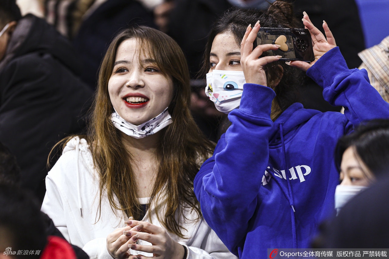 这位女球迷在人群中显得格外明艳，她穿着浅色外套，对着镜头露出自信而大方的微笑，背景里模糊的赛场光影，成了她这幅“冬日观赛图”最好的衬托。