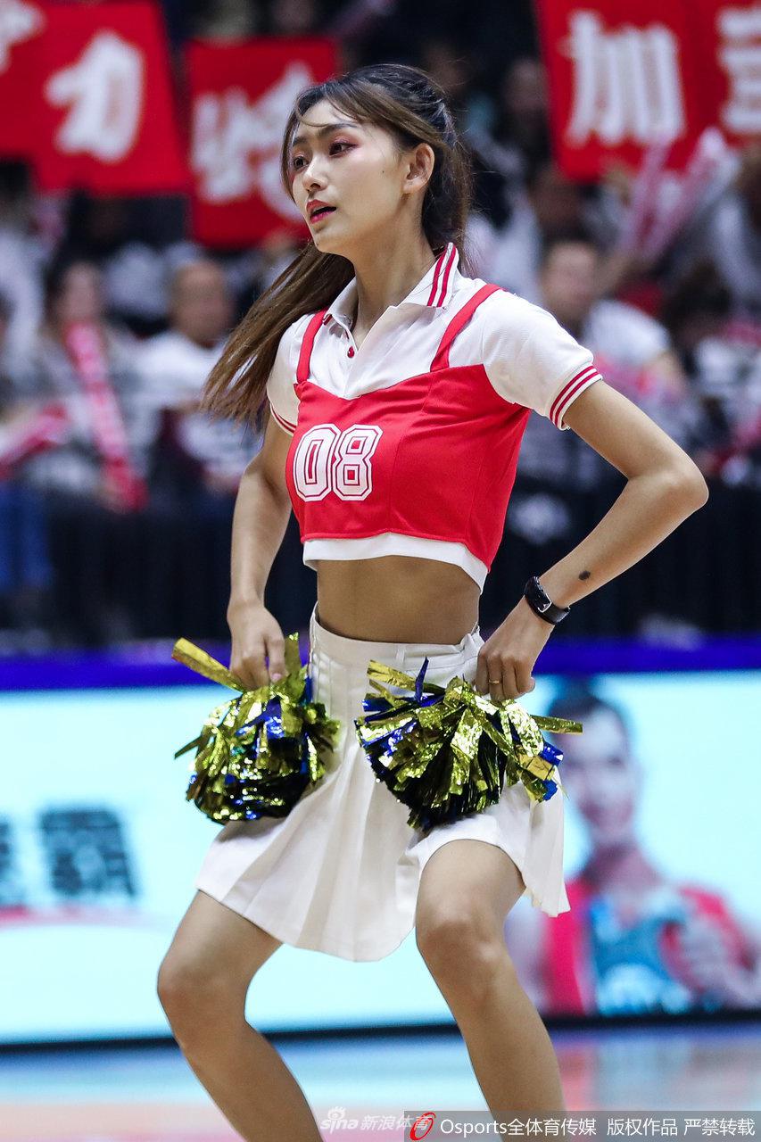 镜头拉近,一位篮球宝贝正在专注地完成舞蹈动作,她精致的五官和自信甜美的笑容极具感染力,身后虚化的看台和队友身影更突显了她的舞台魅力。