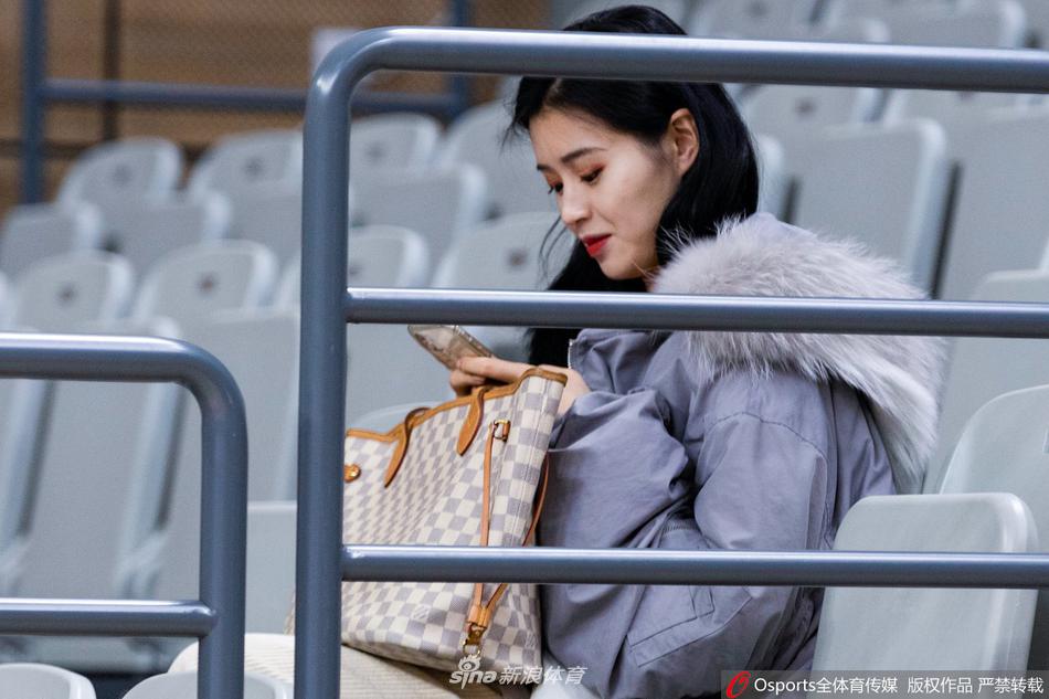 一位穿着休闲卫衣的可爱女球迷，双手捧着手机，背景是模糊的赛场和助威的人群，她或许在记录这一刻，或许在与人分享此刻紧张的心情，青春的气息扑面而来。