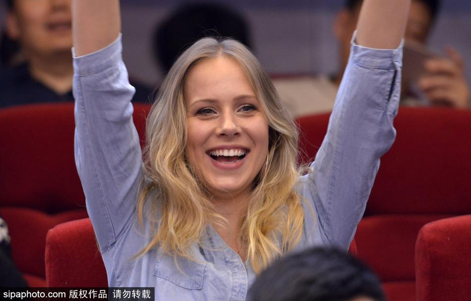 宝莲娜在看台上开心地玩着手机，脸上带着轻松的笑意，身后的球场灯光与观众身影虚化，衬托出她作为场边一道独特风景的悠然自得。