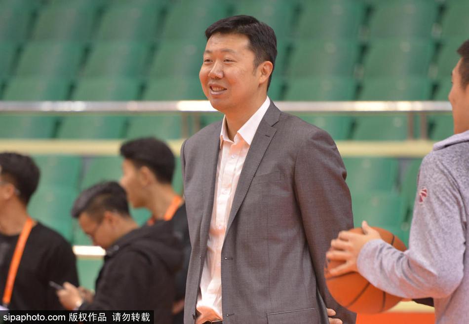 王治郅身穿黑色羽绒服站在赛场中央，双手插兜，面带微笑地注视着远处正在热身的八一队球员，眼神中充满了鼓励与期待。