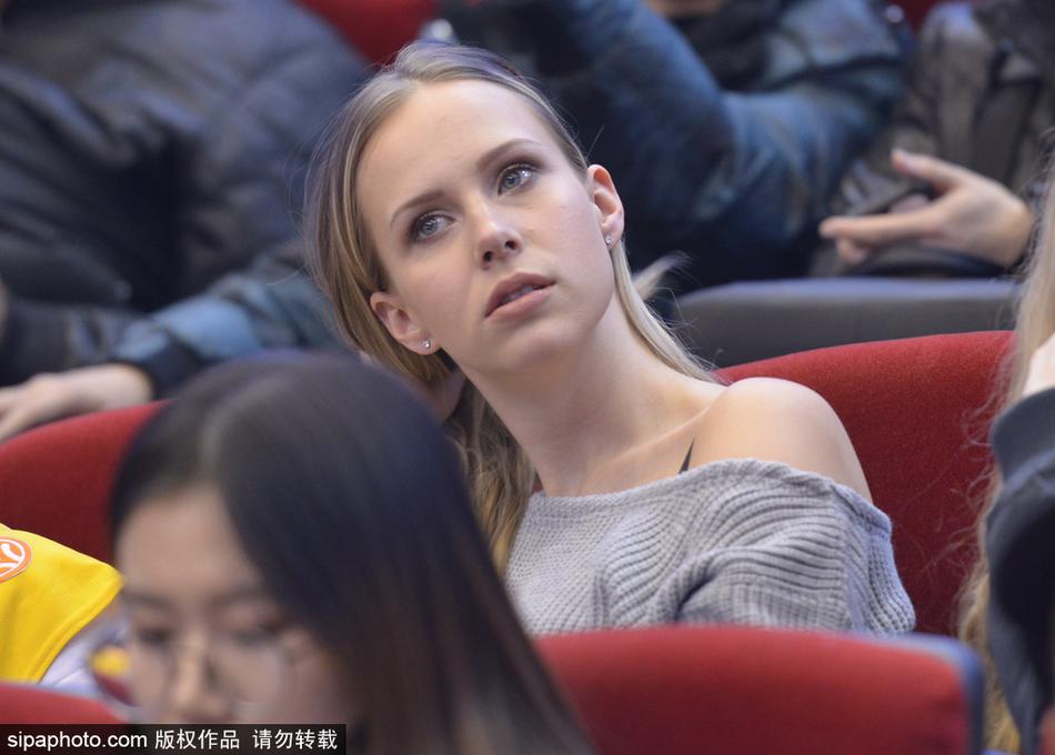 金发飘逸的莫泰妻子身着一袭深色休闲装,坐在看台上露出明媚笑容,仿佛冬日里的一抹暖阳照耀着球场