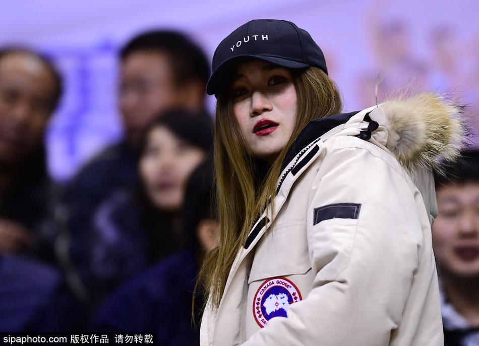 一位长发披肩的美女球迷坐在看台上,她身姿挺拔,穿着时尚的黑色外套,正全神贯注地望向赛场方向,眼神中带着对主队辽宁男篮的无限关切,仿佛下一秒就要为球员的精彩表现起身呐喊。