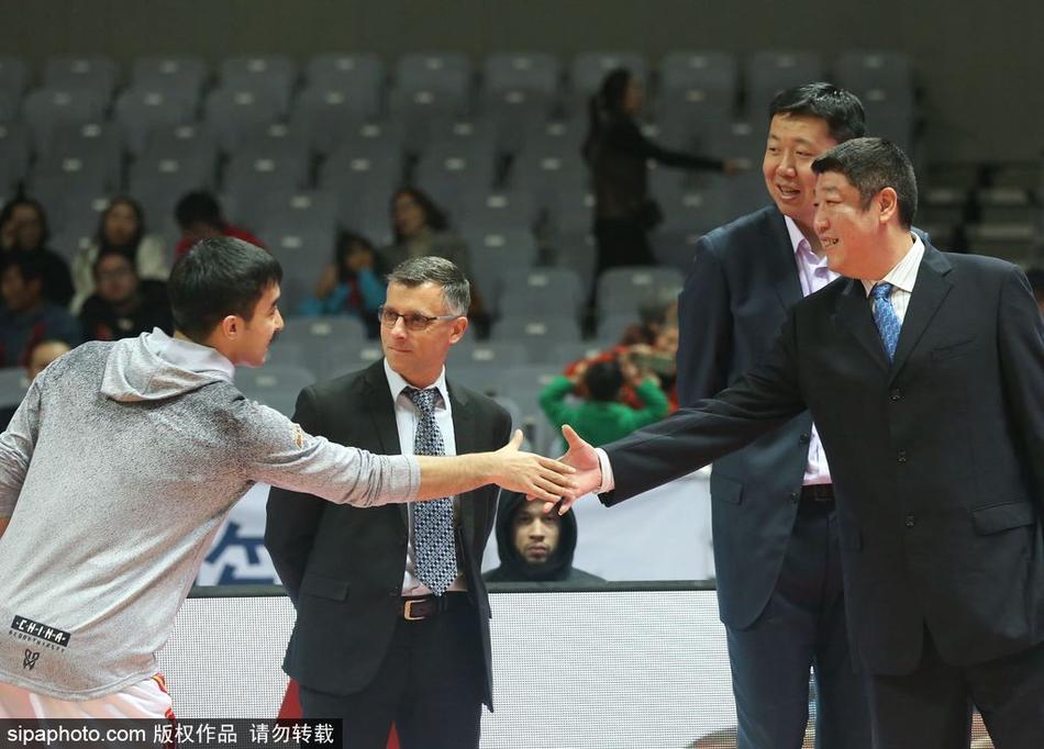 王治郅双手背后，神情专注地注视着场上的八一队年轻队员，身后是正在热身的对手，他的身影显得沉稳而坚定，宛如定海神针。