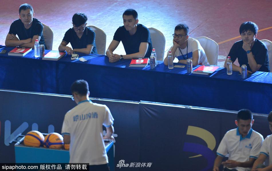 杨鸣坐在评委席上，手拿打分表，神情专注而认真。他身后的背景是热闹的三人篮球赛场，彩旗飘扬，而他仿佛隔绝了周围的喧嚣，沉浸在专业的评判之中。