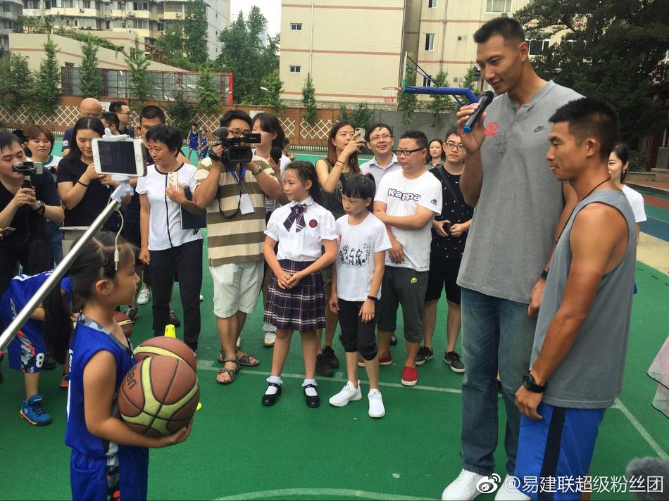 几位小面试官坐在课桌后神情专注,其中一位戴着红领巾的男孩正拿着话筒向站在对面的易建联提问。易建联双手背在身后,微微俯身侧耳倾听,仿佛真的在认真接受一场工作面试。