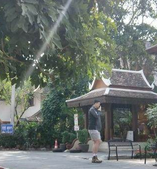 姚明一家在游览间隙停下脚步休息。姚明双手插兜，面带微笑地看向镜头，身后的绿植和泰式建筑衬托出度假的轻松氛围，尽管穿着朴素，但他那挺拔的身姿和亲和的笑容依然是画面中绝对的中心。