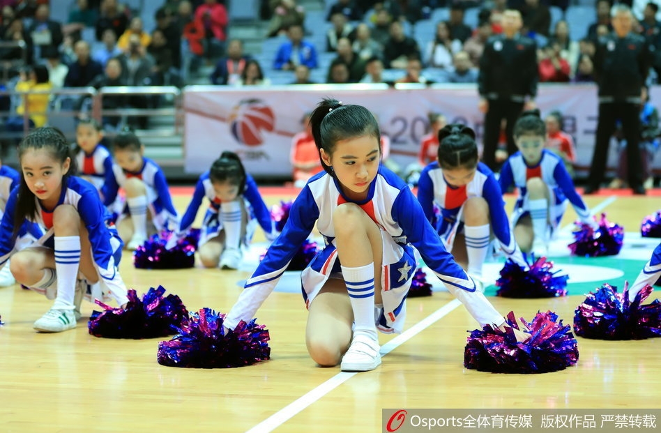 几位扎着丸子头的小宝贝身穿红色球衣，手拿金色花球，踮起脚尖认真观看比赛，眼神里满是好奇与兴奋