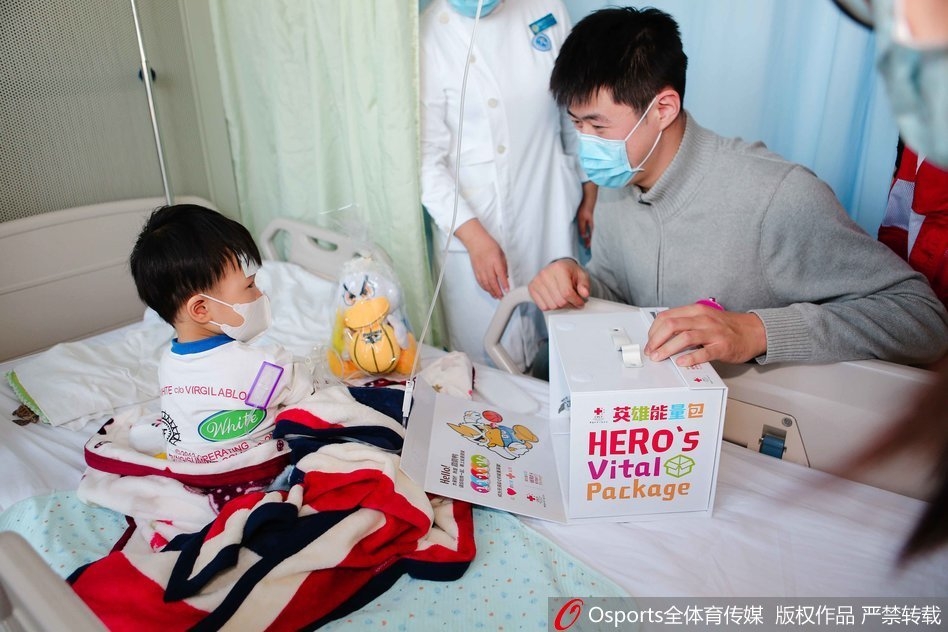 翟晓川、张松涛和吉祥物霹雳鸭一起,手捧着装满礼物的英雄能量包,准备走进病房送给孩子们,他们的脸上洋溢着温暖而真诚的微笑,背景是贴满卡通图案的医院走廊