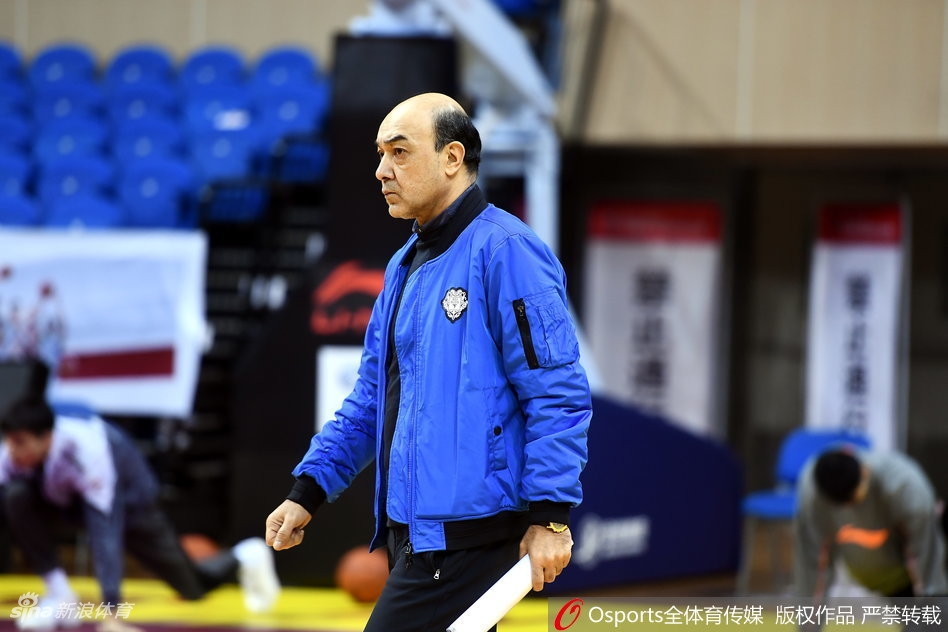 训练间隙,阿的江指导双手叉腰,神情略显疲惫却依然坚毅,他的背后是刻苦训练的年轻队员,一老一少构成了一幅传承的画面。