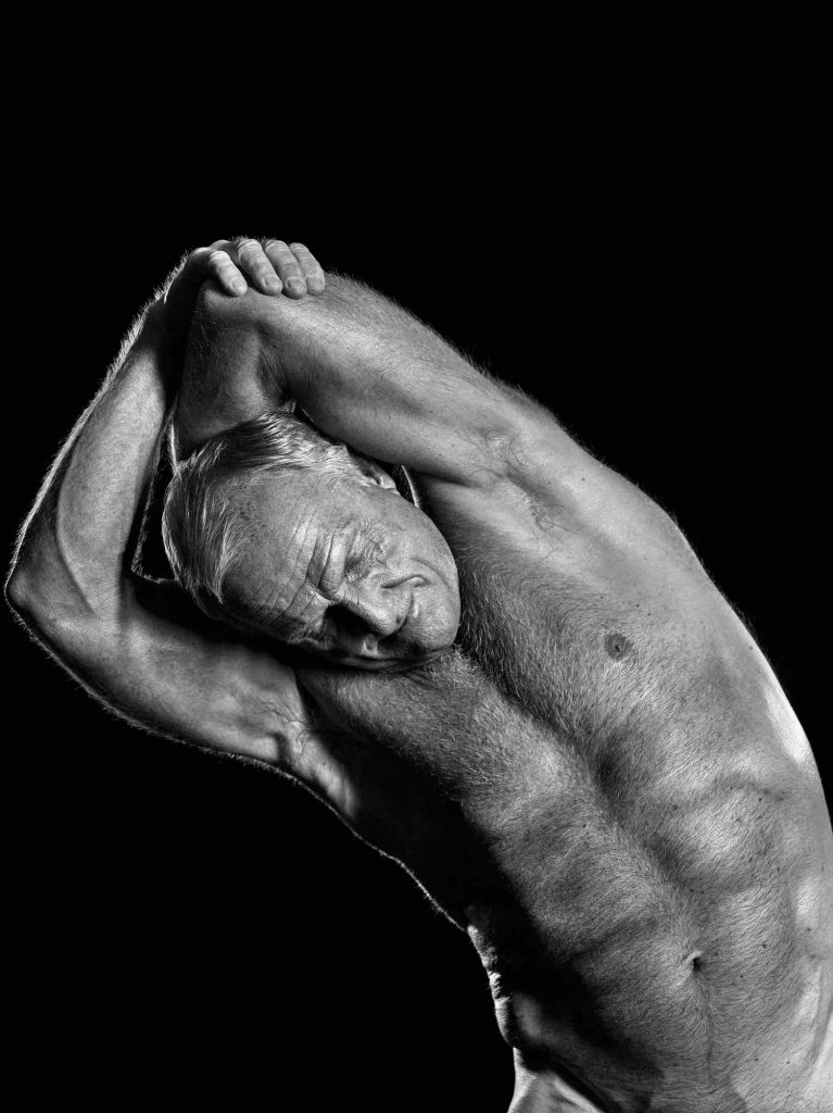 诺曼双臂环抱矗立于礁石之上，背景是澎湃的海浪，整体构图充满古典英雄雕塑般的力量与美感。