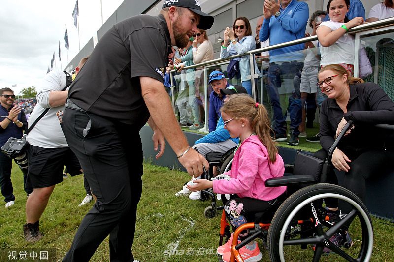 肖恩-劳瑞弯下腰将签名高尔夫球递给围绳外伸手期待的小球迷，孩子脸上洋溢的惊喜与球员温和的笑容温暖了整个赛场