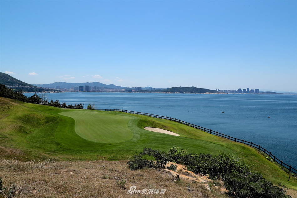 从高处俯瞰球场一角，宽阔的果岭如同镶嵌在海岸线上的翡翠，被蜿蜒的球道和湛蓝的海水环绕，景色壮丽非凡。