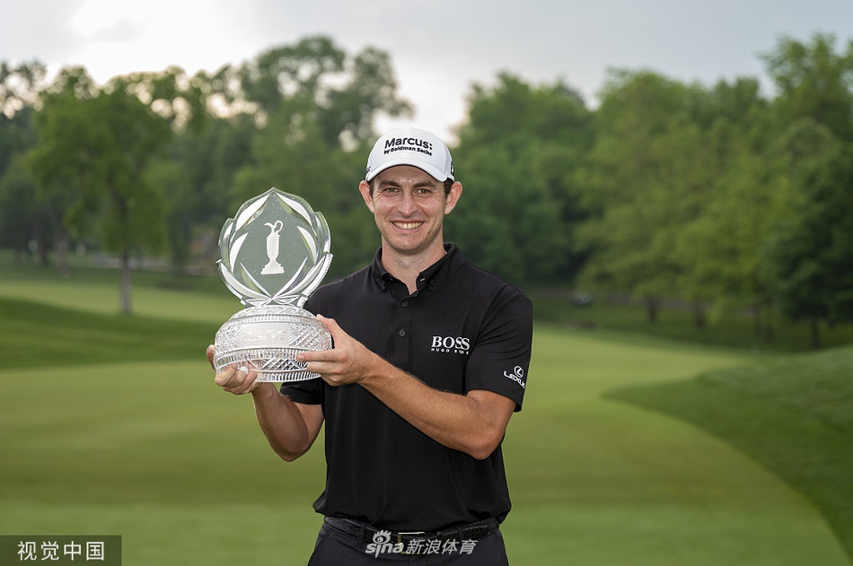 帕特里克·坎特利高举纪念高球赛冠军奖杯，笑容灿烂，身后是绿意盎然的缪菲尔德山庄和为他欢呼的观众。