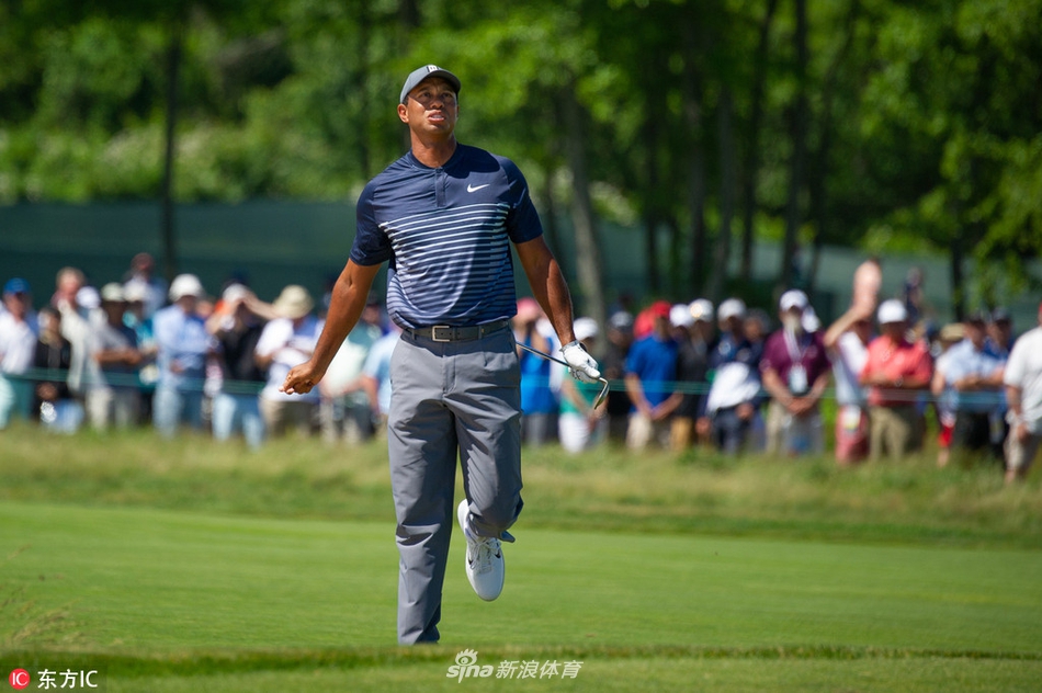 伍兹扛着球包行走在沙坑与长草间，背景是广阔的球场，他的身影显得孤独而坚定。