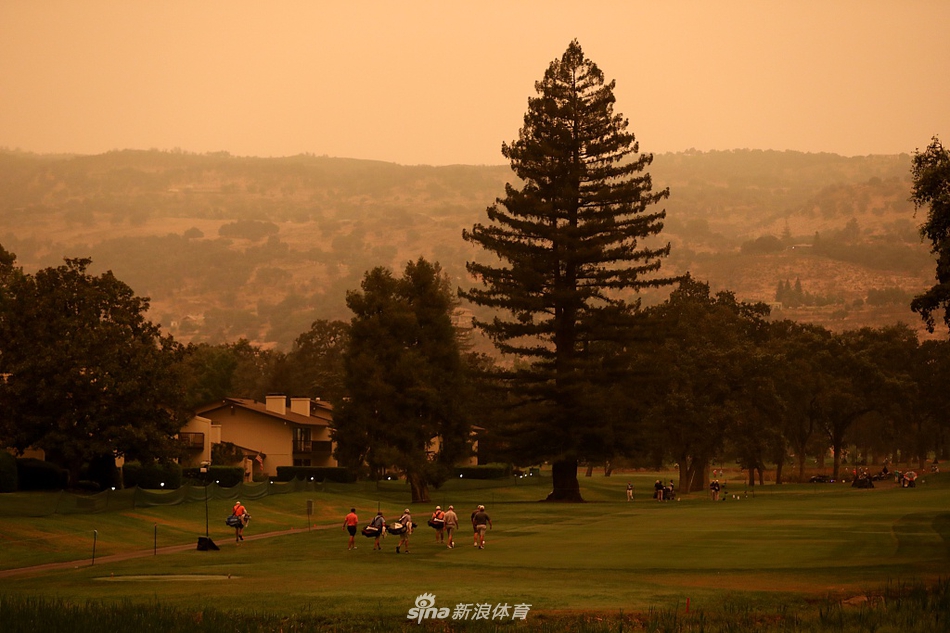 一位球童背着硕大的高尔夫球包走在球道上，他的剪影投射在橙红色的朦胧光晕中，象征着赛事在巨大自然力量面前依然稳步前行。