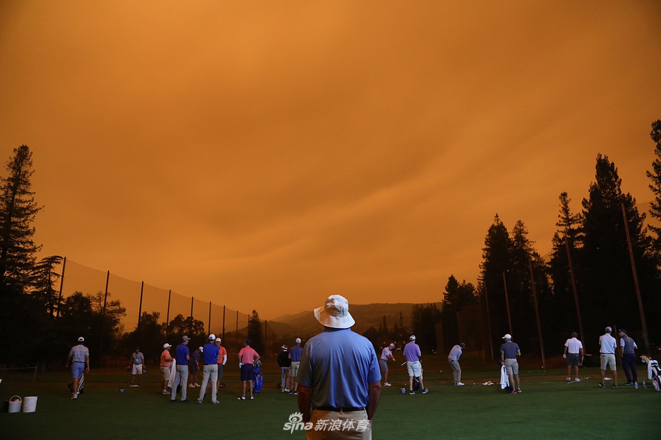 赛事工作人员在发球台附近进行准备工作，他们身后，练习果岭的旗帜在橙红色的天空映衬下静静垂下，等待比赛开始。