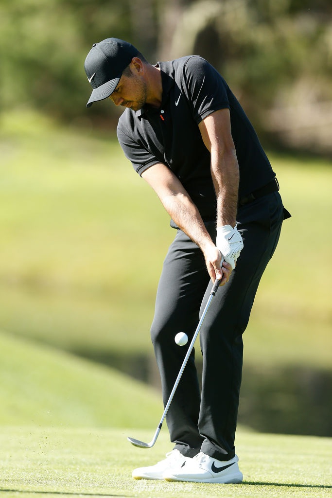 简森·戴伊在练习果岭边进行切杆练习，阳光洒在他的身上，动作优雅而稳定