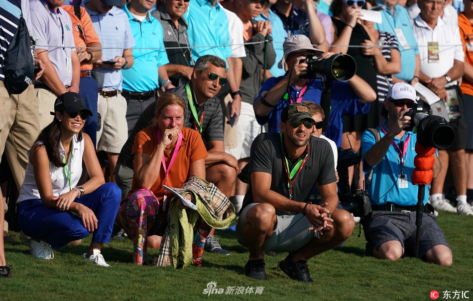 菲尔普斯独自站在球场围绳边，双手插在裤袋中，微微侧身凝视着果岭方向。阳光洒下，他的表情沉静而若有所思，完全沉浸在高尔夫赛事的氛围里。