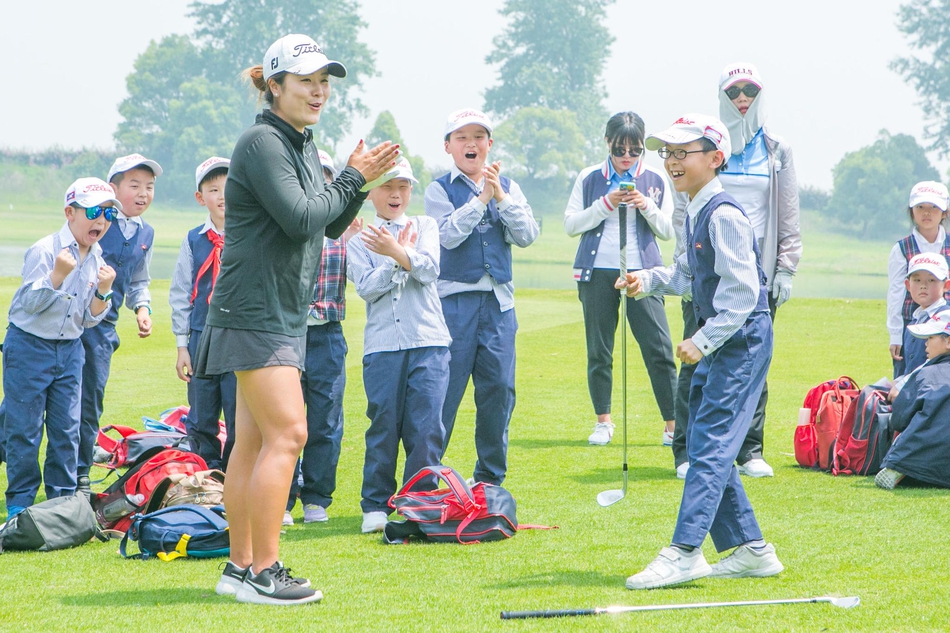 杰奎琳正俯身悉心指导一位小球迷，两人一同握杆，专注调整挥杆姿势