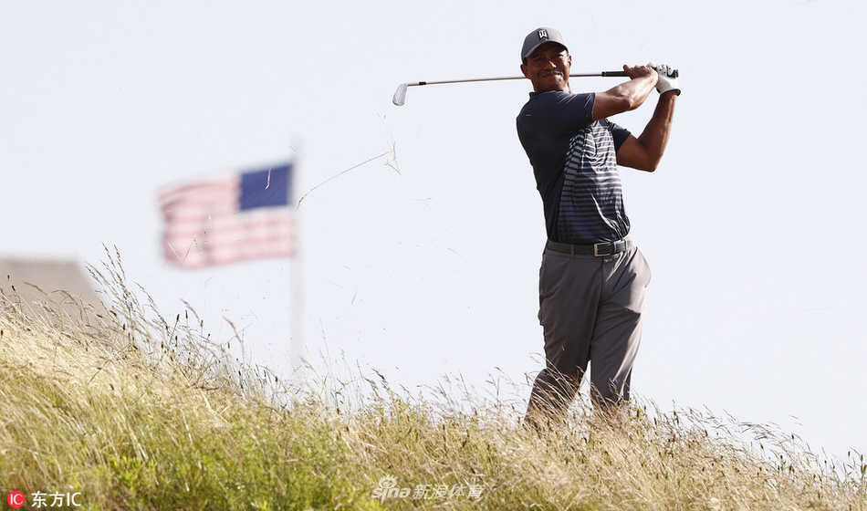 伍兹站在发球台旁，双手扶膝，面色凝重地观察着前方球道，强风吹动了他的衣角和头发。