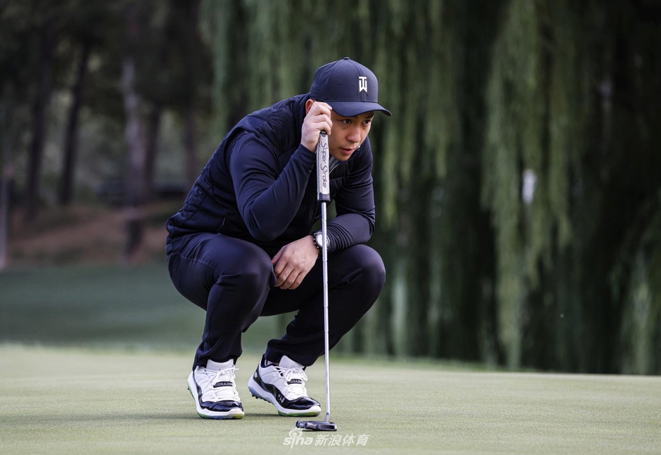 释小龙半蹲在果岭边，神情极为专注地观察球路与草纹，用手细致地比划推杆线路，尽显沉稳与耐心。