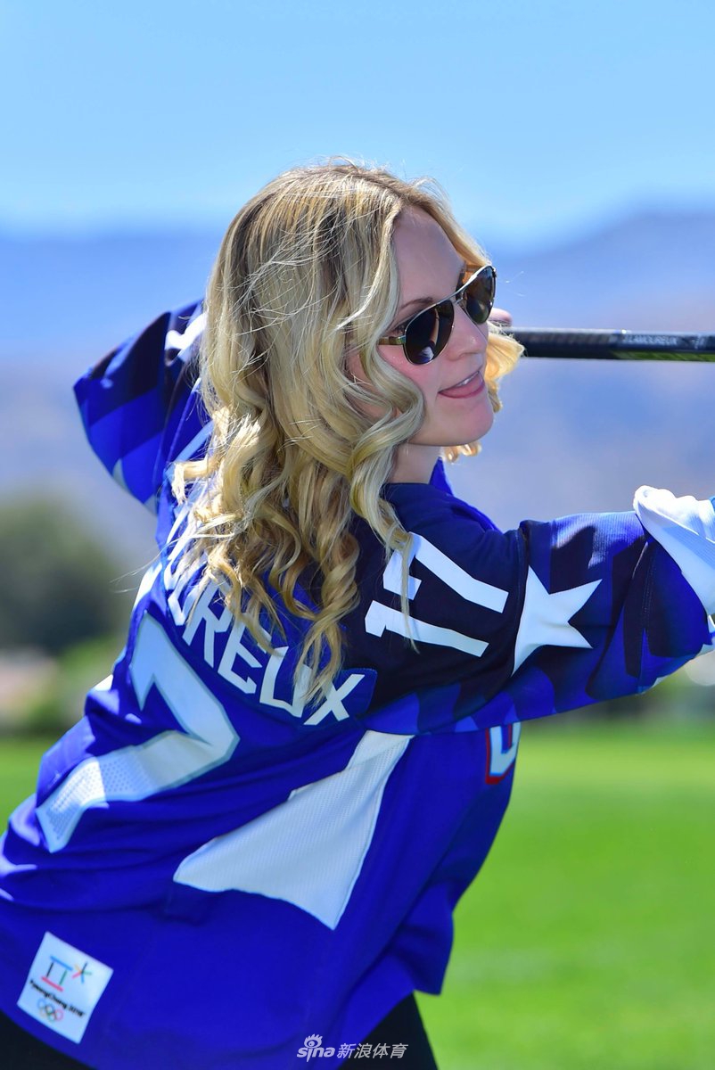 好莱坞影星艾什莉·贾德手持高尔夫球，面向镜头露出标志性温暖笑容，身后是广阔的练习果岭。