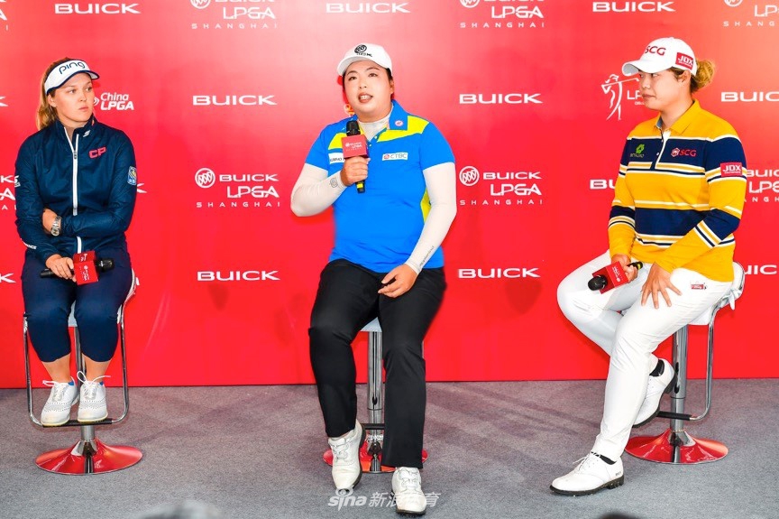 赛场全景风貌：阳光下的高尔夫球场绿意盎然，蜿蜒的球道、清澈的水塘与起伏的沙坑构成一幅壮丽画卷，球员与观众点缀其中。