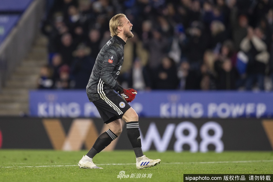 小舒梅切尔在球门前冷静观察场上局势，身体微微下蹲，随时准备做出扑救反应，球门后的看台上是模糊的球迷身影。