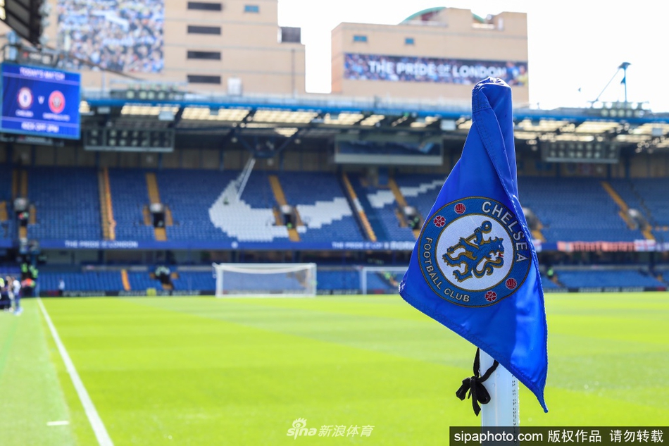 一位NHS医护人员疲惫却坚定地站在酒店房间内，窗外是熟悉的斯坦福桥球场一角，这方小小的空间成为了她们在抗疫风暴中可以安心停泊的宁静港湾。