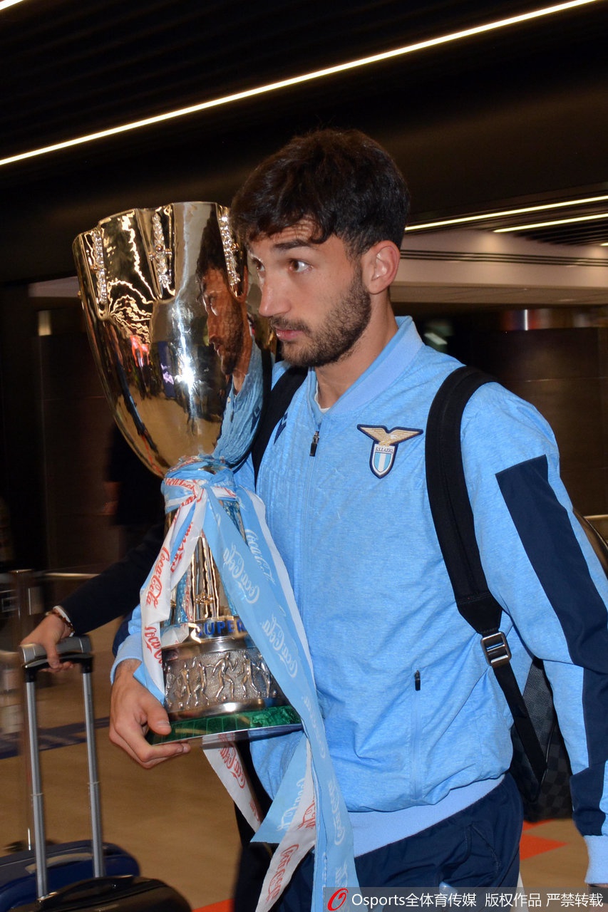 在球迷的簇拥中，一名年轻的拉齐奥小球迷眼神中充满了对偶像的崇拜与向往，他高举着手机，试图记录下这难忘的瞬间，背景是喧嚣而喜庆的人群。